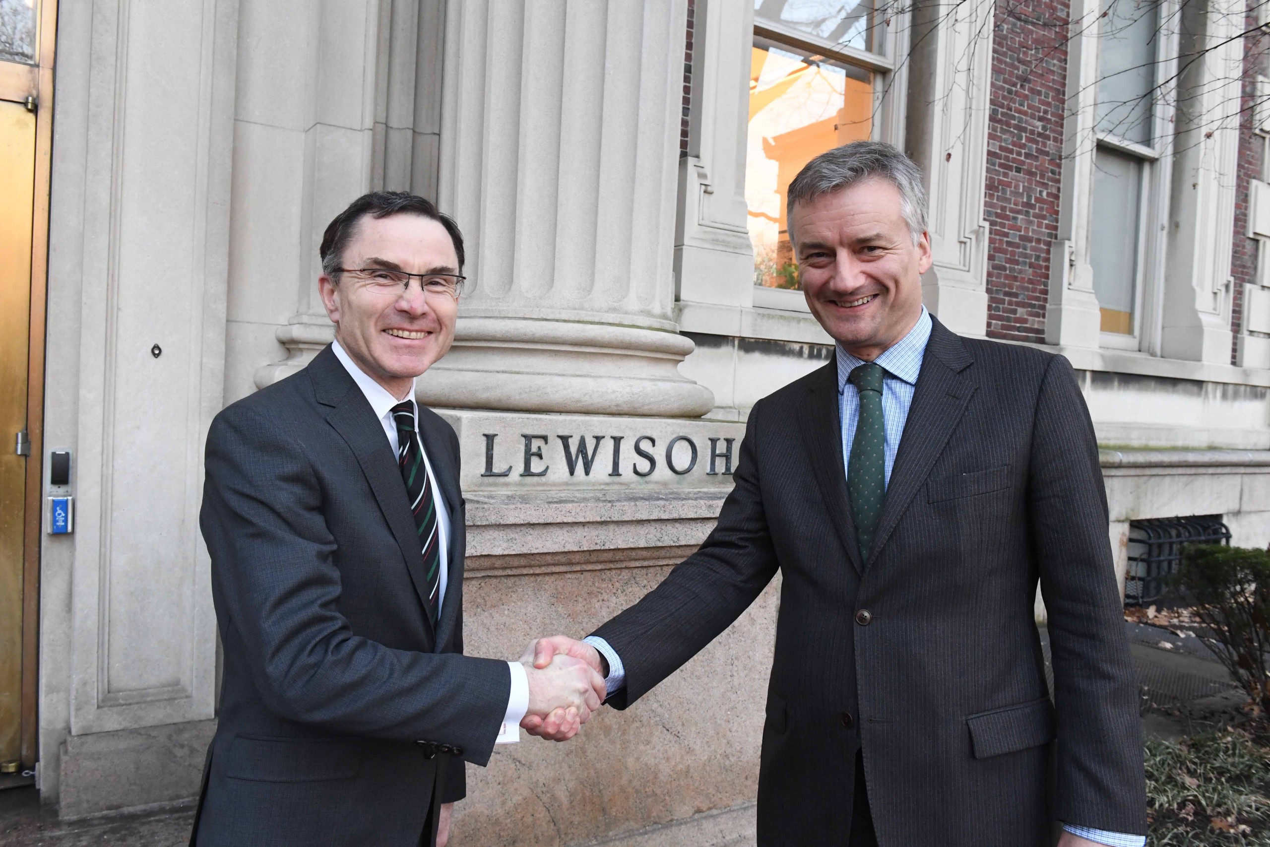 Two men in suits smile at the camera while shaking hands